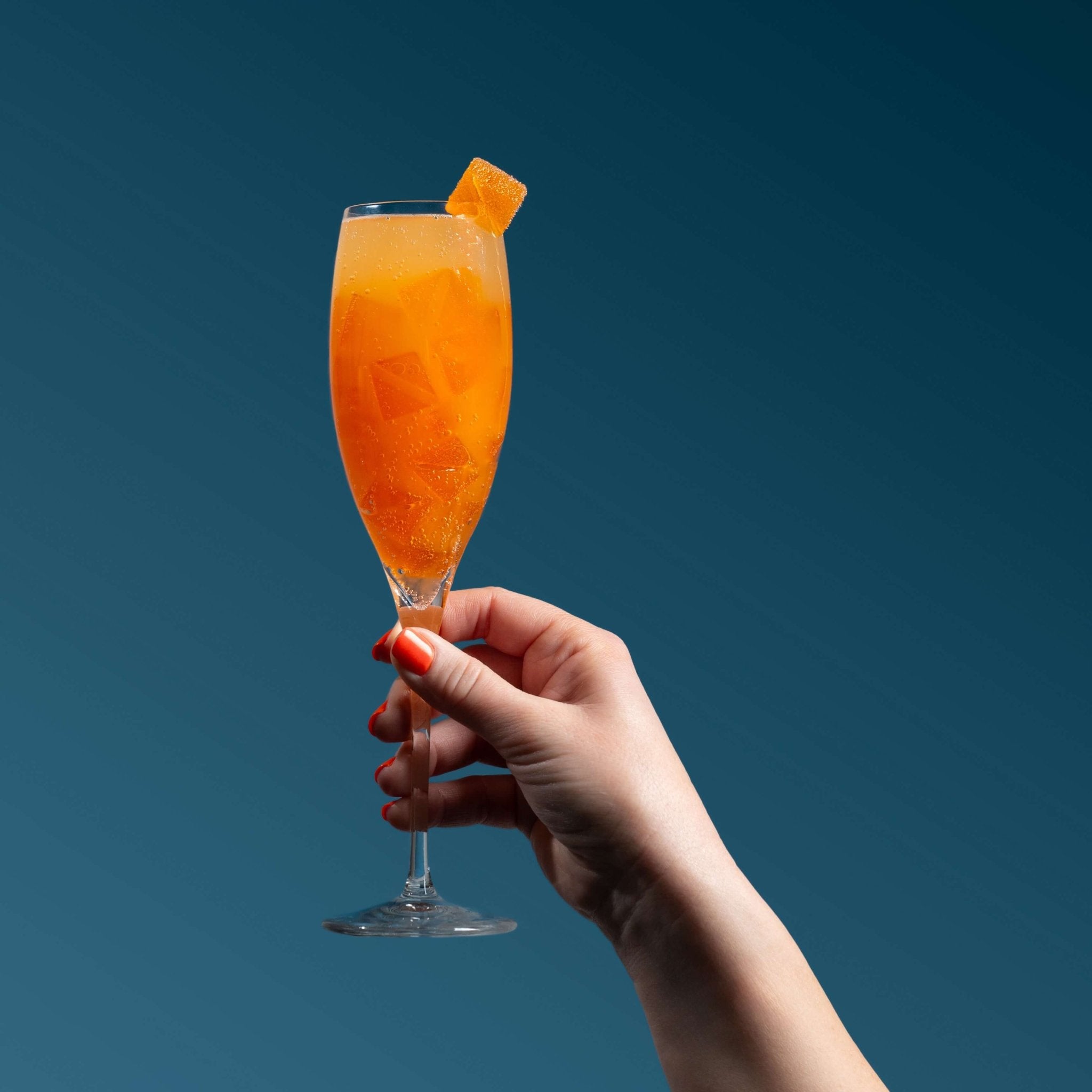 Nothing But Hemp Mimosa hemp-derived THC and CBD sativa gummies shown in a champagne glass filled with orange gummies and champagne, with a gummy garnished on the rim, held by a hand against a dark blue background in a lifestyle scene